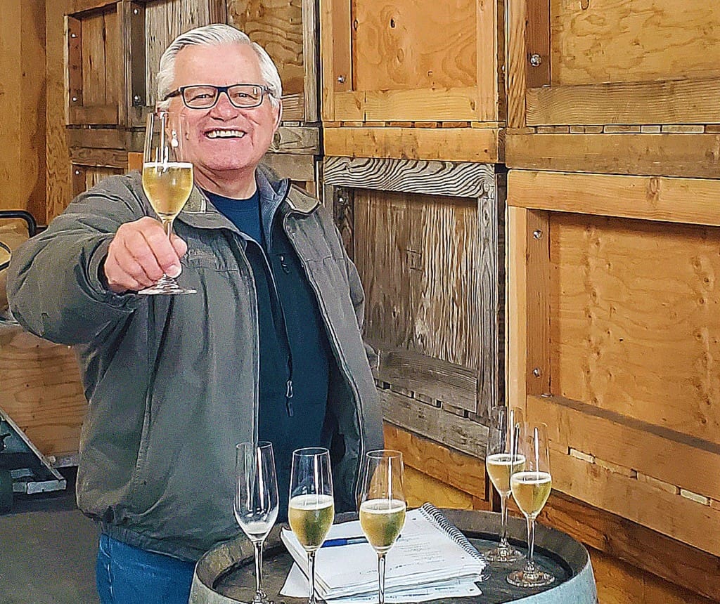 Smiling man holding a champagne glass, standing by a barrel with more glasses and a notebook on top.