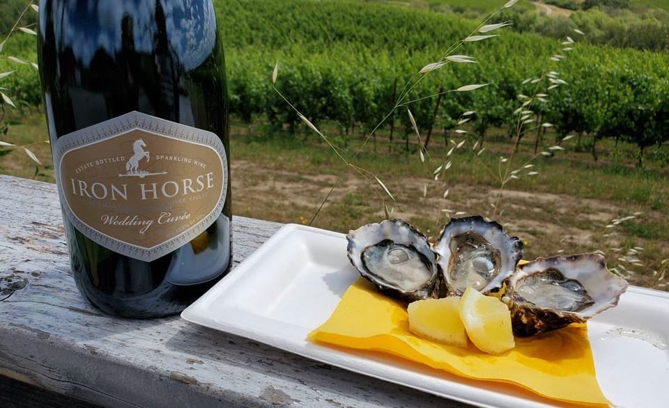 Bottle of Iron Horse sparkling wine next to oysters and lemon wedges on a plate, with vineyard in background.