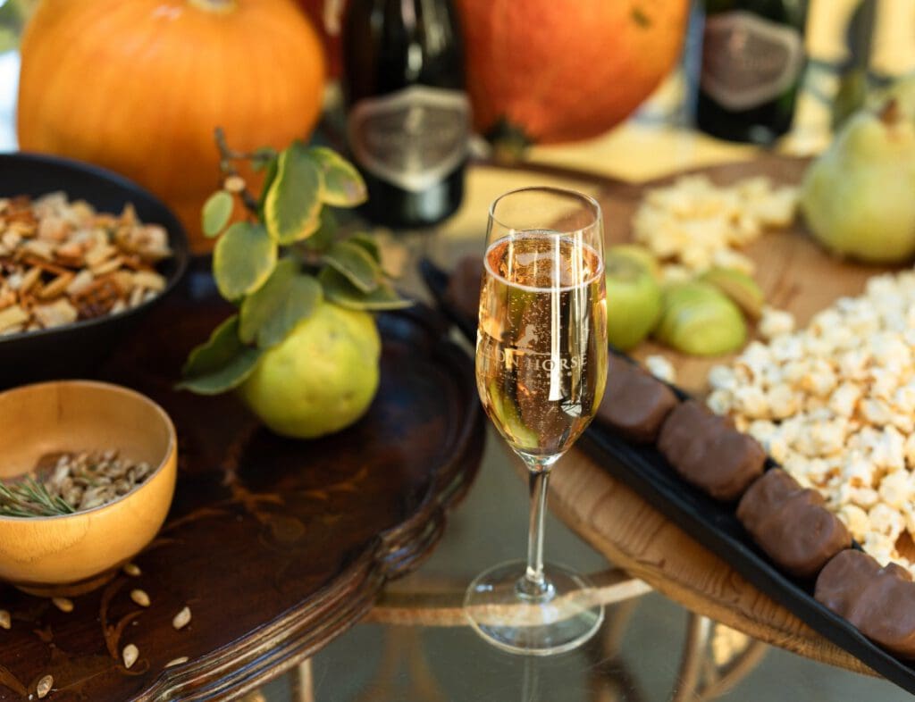 A glass of sparkling wine on a table with popcorn, chocolate bars, fruit, seeds, and pumpkins.