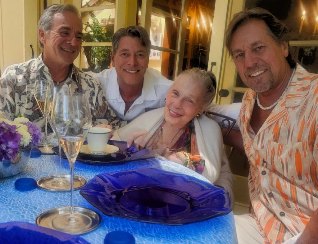 Four smiling adults sit around a table with blue plates, flowers, and drinks at an outdoor gathering.