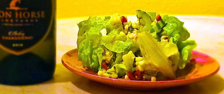 A lettuce salad with corn and red peppers on a plate, next to a dark bottle, against a yellow background.