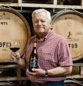Older man in a plaid shirt holding a wine glass and bottle, standing in front of wine barrels.
