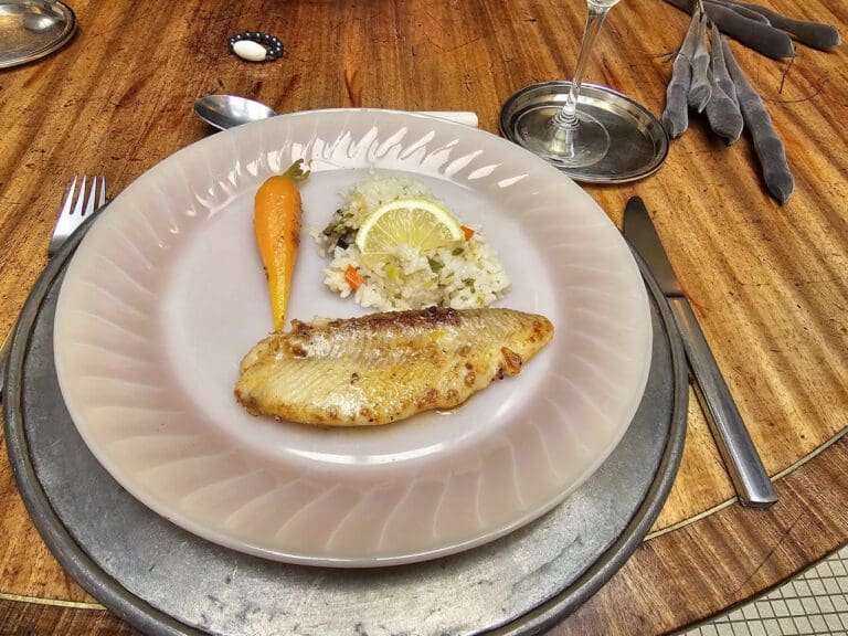 A plate with grilled fish, rice with lemon, and a cooked carrot on a wooden table.