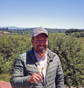 Man in a gray cap holding a glass outdoors, with green trees and hills in the background.