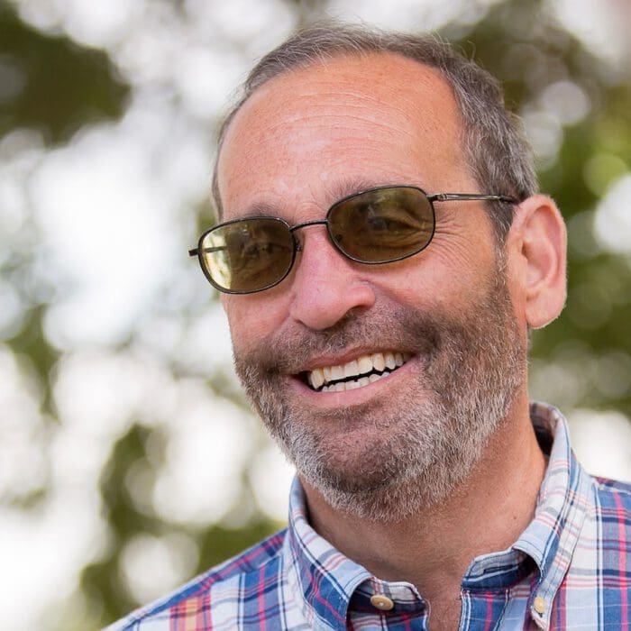 Smiling man with a beard and sunglasses wearing a plaid shirt, outdoors with trees in the background.