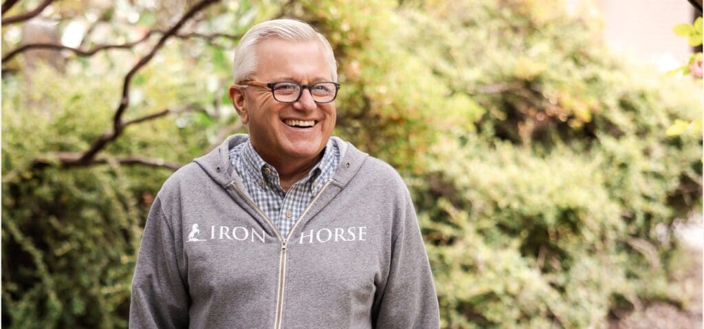 Smiling man in glasses and a gray Iron Horse hoodie stands outside with greenery in the background.