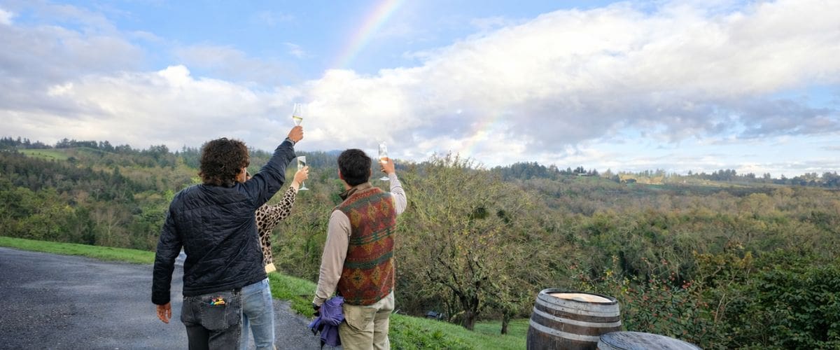 catching a rainbow in a sparkling wine glass Three people raise glasses toward a rainbow over a scenic, green landscape with a barrel in the foreground.