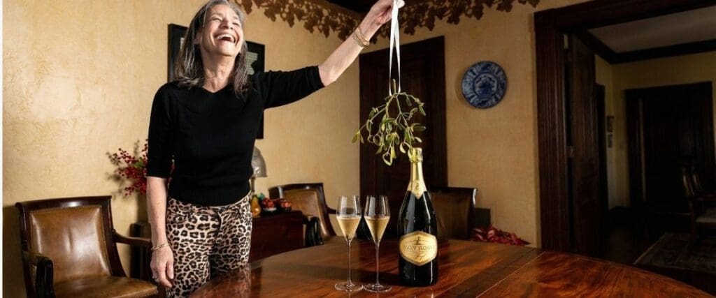 A woman smiles, holding mistletoe over a table with champagne and glasses in a warmly lit room.