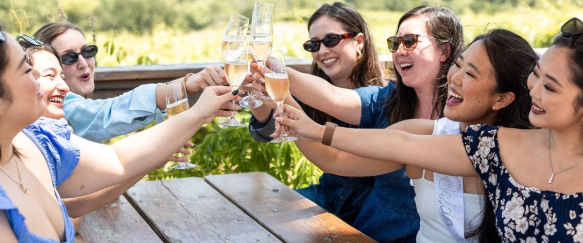 ladies toasting outside Seven women smiling and clinking glasses of champagne outdoors at a wooden table on a sunny day.