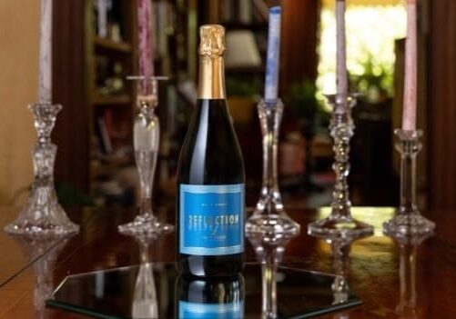 A bottle of sparkling wine labeled Reflection on a mirrored surface, surrounded by lit candles.