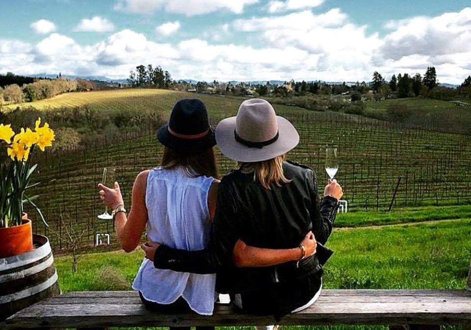 Two people in hats sit on a bench, overlooking a vineyard and holding wine glasses, with flowers nearby.