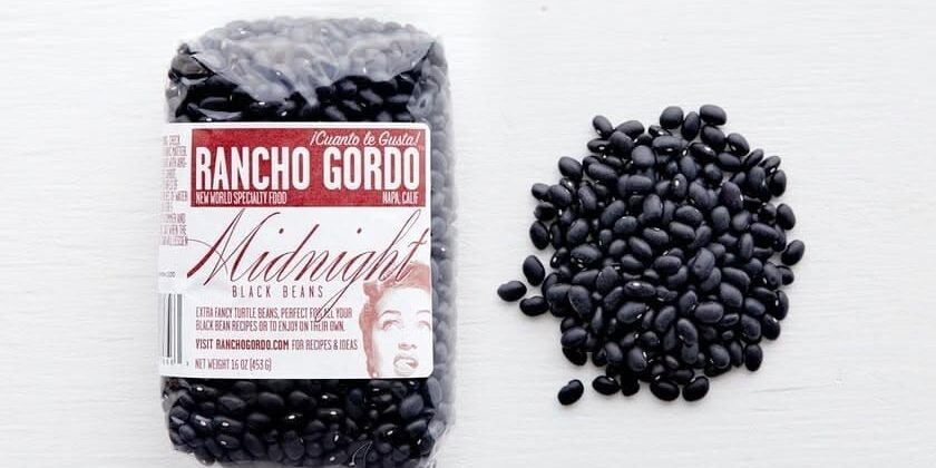Black-Beans A bag of Rancho Gordo Midnight black beans next to a pile of loose black beans on a white surface.