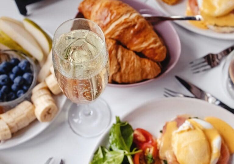 A glass of sparkling wine with croissants, eggs benedict, fruit, and greens on a breakfast table.