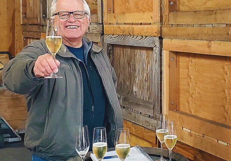 Smiling man holding a champagne glass, standing by a barrel with more glasses and a notebook on top.