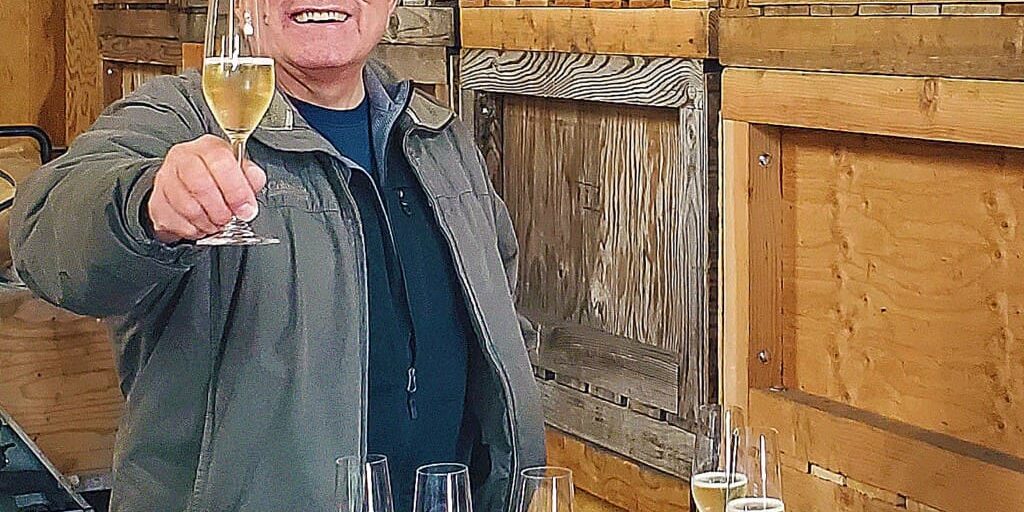 Smiling man holding a champagne glass, standing by a barrel with more glasses and a notebook on top.