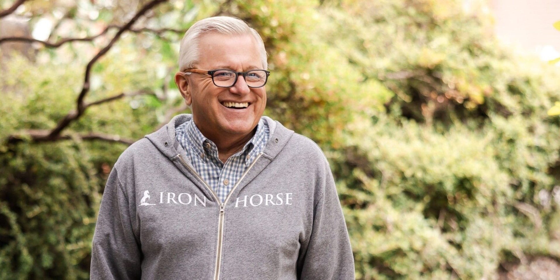 Smiling man in glasses and a gray Iron Horse hoodie stands outside with greenery in the background.