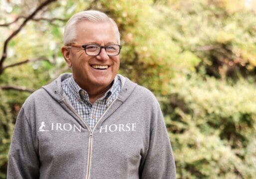 Smiling man in glasses and a gray Iron Horse hoodie stands outside with greenery in the background.