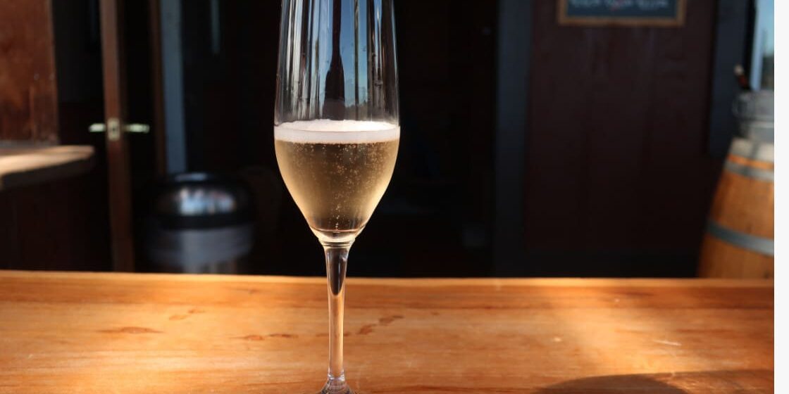 A glass of sparkling wine on a wooden bar with a dark background and light shining on the glass.