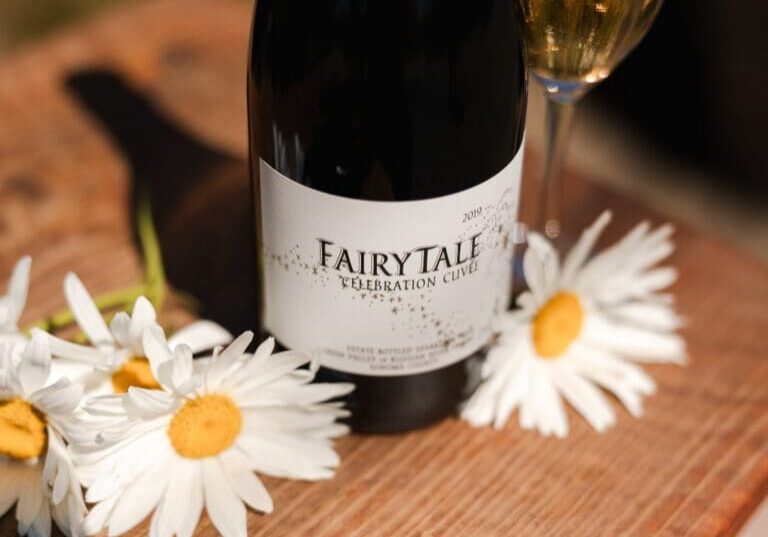 A bottle of Fairytale Celebration Cuvee wine with daisies and a filled wine glass on a wooden surface.