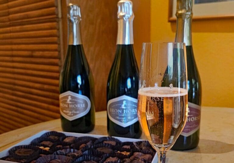 Three bottles of sparkling wine, a glass of rosé, and a box of assorted chocolates on a table.