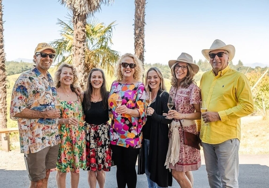 Seven smiling adults in colorful outfits stand outdoors, holding drinks, with palm trees and blue sky behind them.
