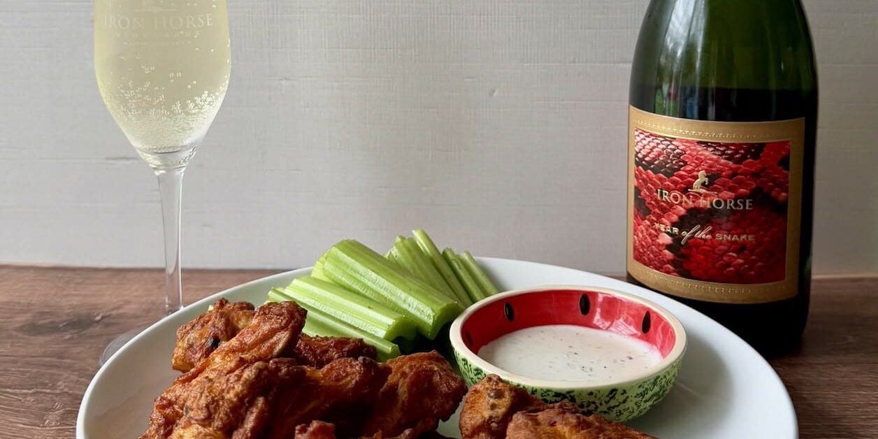 Plate of buffalo wings, celery sticks, and ranch dip with a glass and bottle of sparkling wine on a table.