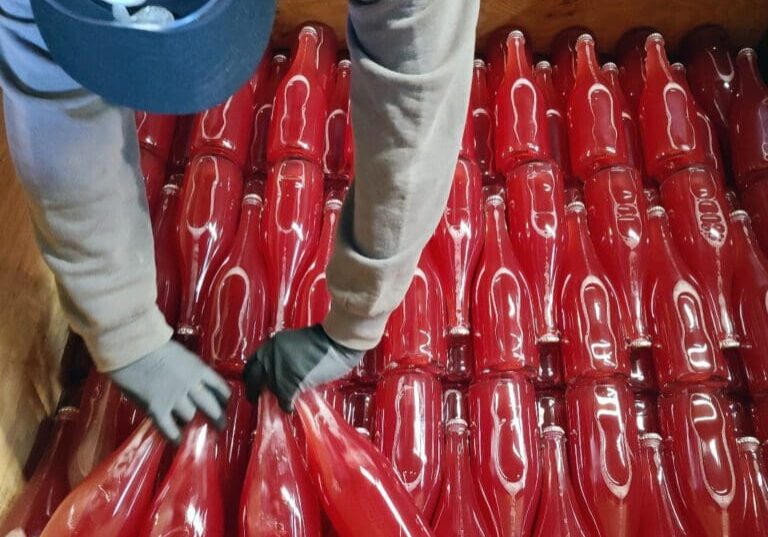 Person wearing gloves arranging red bottles stacked horizontally in a wooden crate.