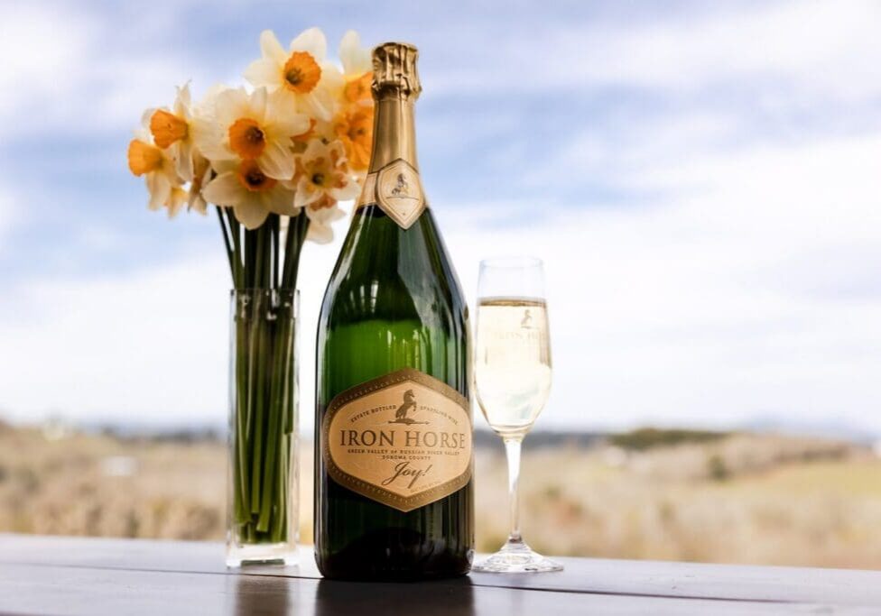 A bottle of Iron Horse champagne, a filled glass, and daffodils in a vase on an outdoor table.
