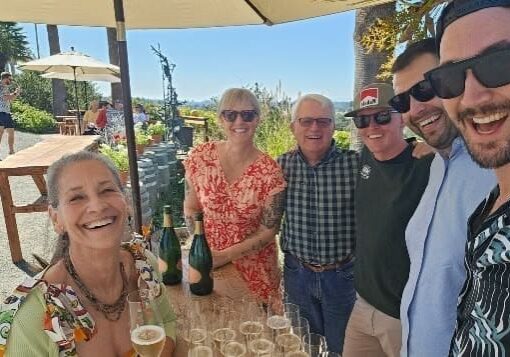 Six smiling people posing outdoors around a table with wine bottles and glasses on a sunny day.