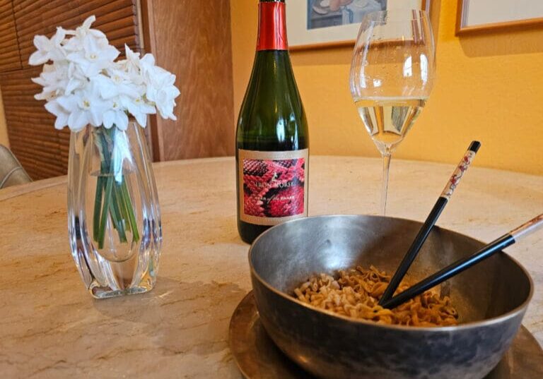 A bowl of noodles with chopsticks, a wine glass, a wine bottle, and white flowers on a table.