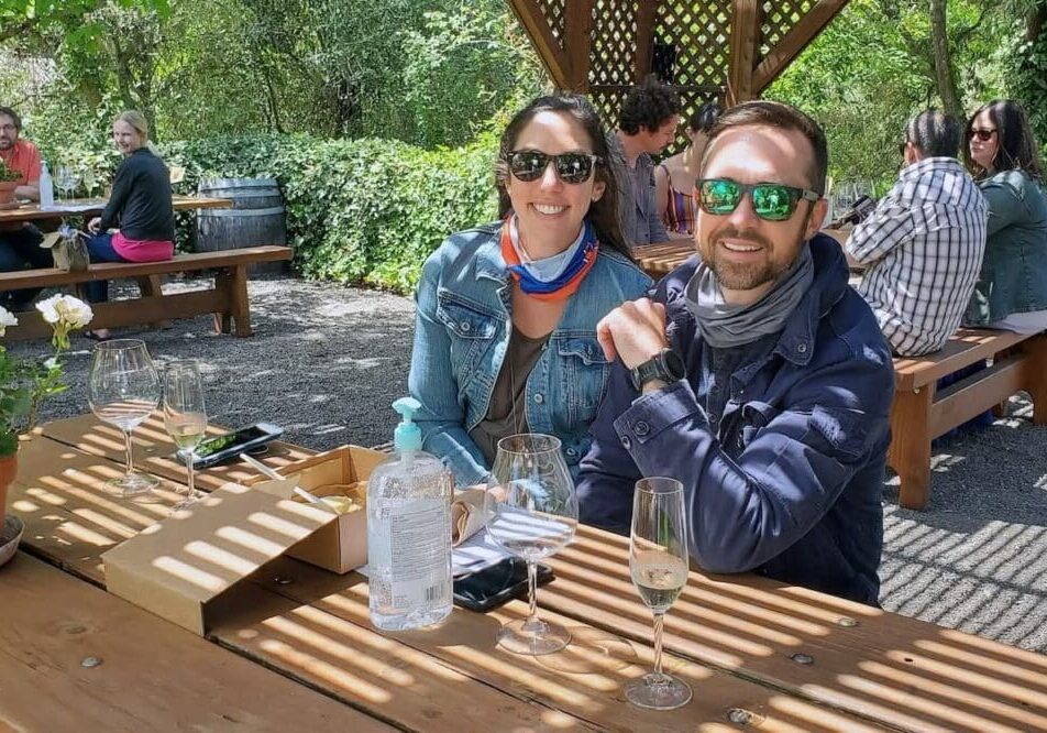 Two people smiling at an outdoor table with wine glasses, wearing sunglasses, on a sunny day.