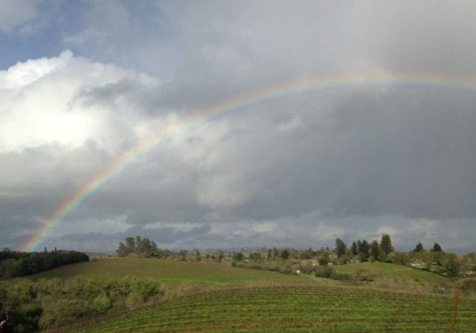 A rainbow arches over a green landscape with hills, trees, and a cloudy sky.