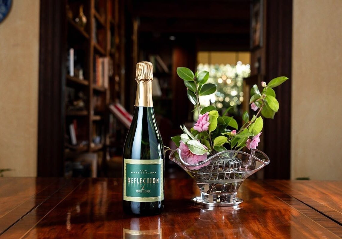 A bottle of sparkling wine and a vase of pink flowers on a polished wooden table indoors.