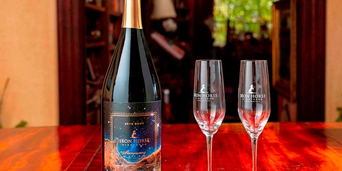 A bottle of Iron Horse sparkling wine and two empty glasses sit on a polished wooden table.