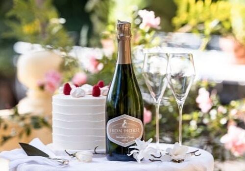 A bottle of champagne, two glasses, and a frosted cake with raspberries on a table outdoors with flowers.