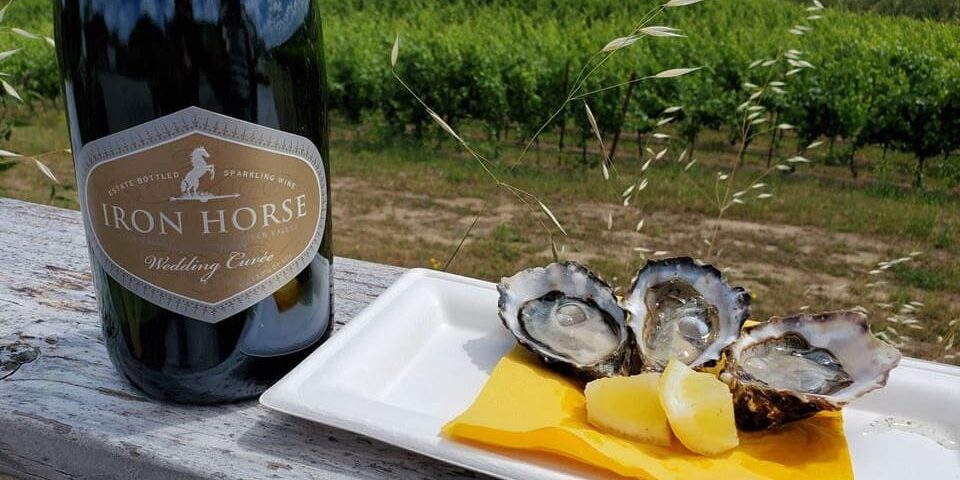 Wedding-Cuvee-Oysters-great-sparkling-wine-pairing Bottle of Iron Horse sparkling wine next to oysters and lemon wedges on a plate, with vineyard in background.