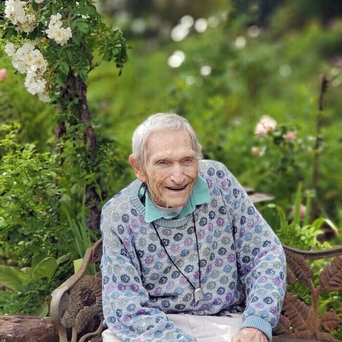 An elderly person with gray hair smiles while sitting on a bench in a lush, flower-filled garden.