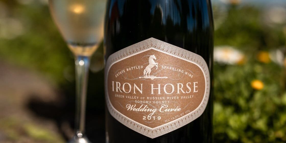 iron-horse-wedding-cuvee-bottle-with-flute A bottle of Iron Horse sparkling wine beside a filled glass, outdoors with blurred greenery and flowers.