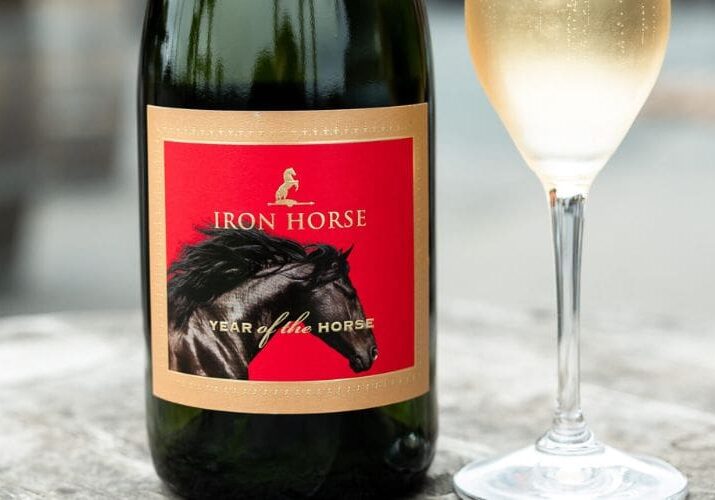 A bottle of Iron Horse sparkling wine with a horse label next to a filled wine glass on a wooden table.
