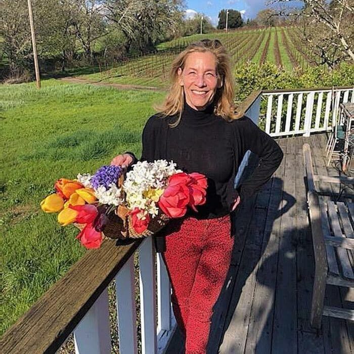 Smiling woman holding a colorful bouquet on a porch overlooking a green field and vineyard.