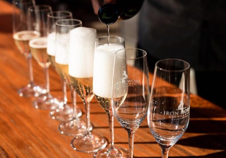Champagne being poured into a row of glasses on a wooden table, some glasses already filled and foaming.