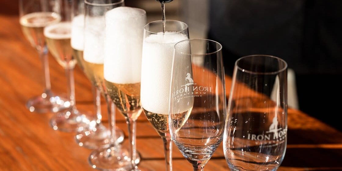Champagne being poured into a row of glasses on a wooden table, some glasses already filled and foaming.