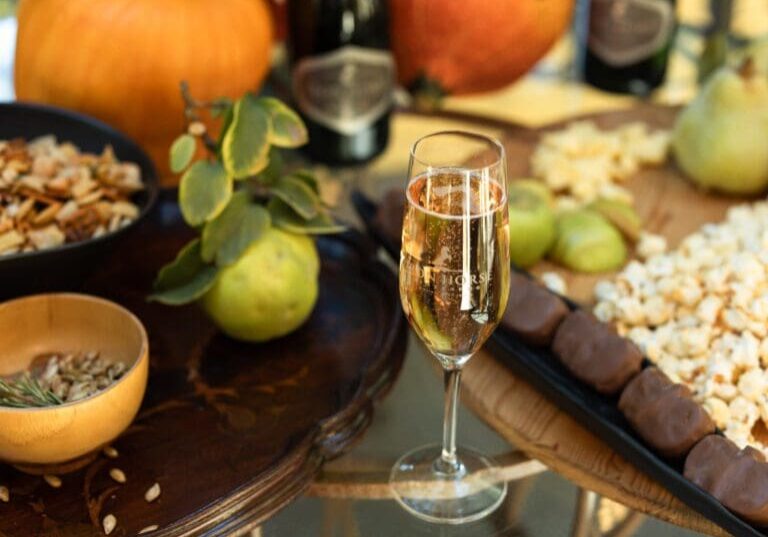 A glass of sparkling wine on a table with popcorn, chocolate bars, fruit, seeds, and pumpkins.