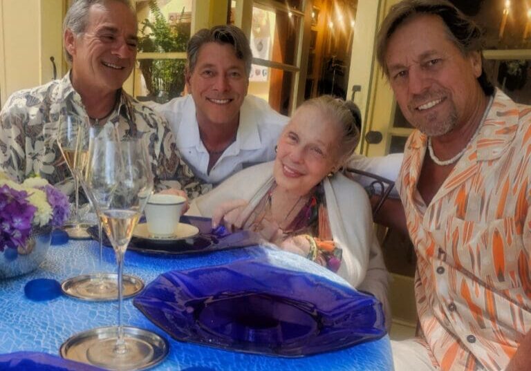 Four smiling adults sit around a table with blue plates, flowers, and drinks at an outdoor gathering.