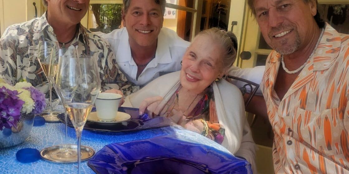 Four smiling adults sit around a table with blue plates, flowers, and drinks at an outdoor gathering.