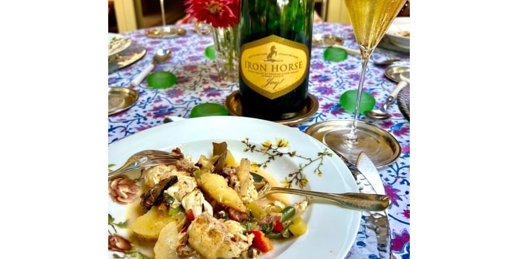 A plate of seafood stew and a glass of sparkling wine beside an Iron Horse wine bottle on a floral tablecloth.