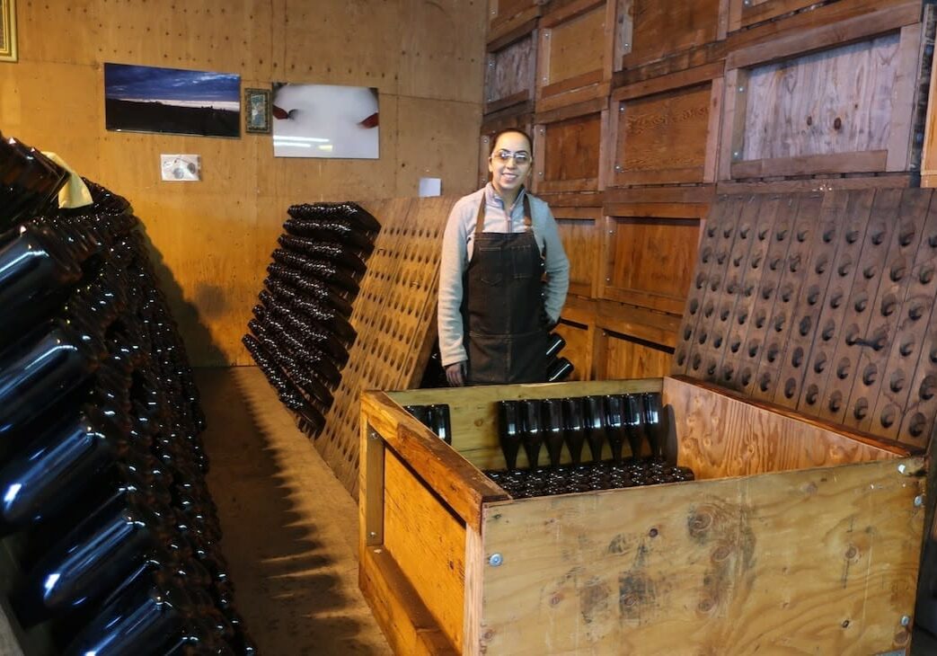 Woman in a winery stands among racks and crates filled with wine bottles in a wooden room.