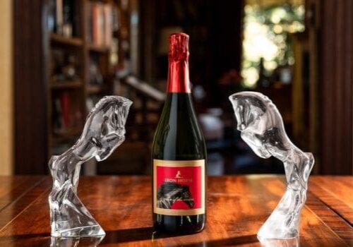 A bottle of Iron Horse sparkling wine stands between two glass horse head sculptures on a wooden table.